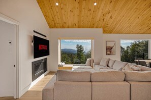 Una sala de estar moderna con un amplio sofá modular, una chimenea, un televisor y ventanas grandes que ofrecen vistas al exterior.