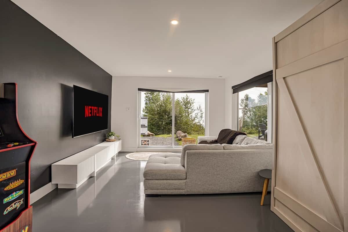 Una sala de estar moderna con un sofá gris, un televisor de pantalla plana montado en la pared y un gran ventanal con vistas al exterior.