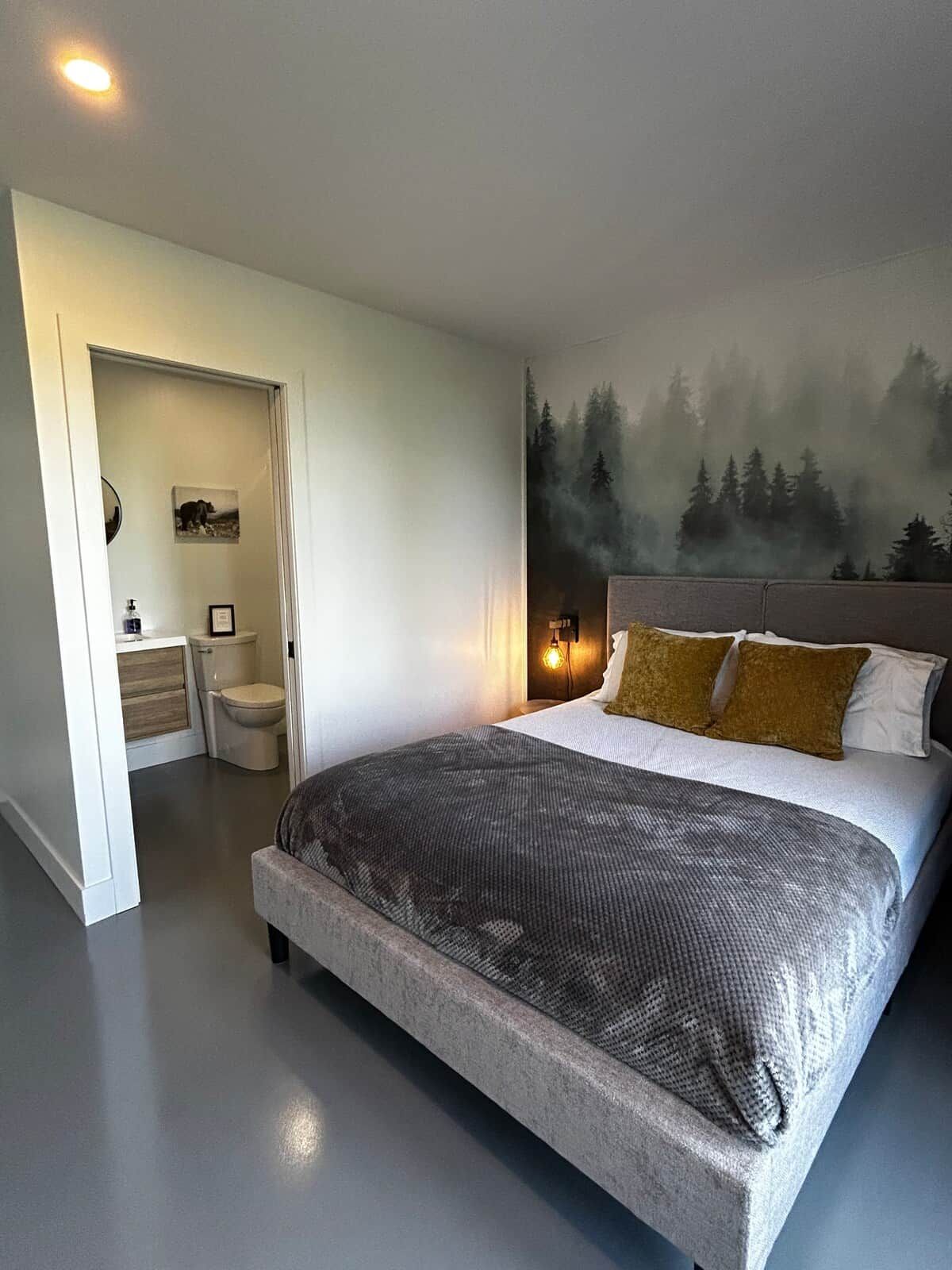 A bedroom with a large bed, a nightstand, a wall-mounted lamp, and a forest mural.