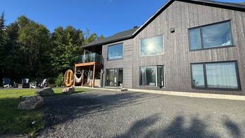 Un bâtiment moderne à deux étages, doté d’une allée en gravier, de grandes fenêtres et d’une terrasse en bois avec des meubles d’extérieur.