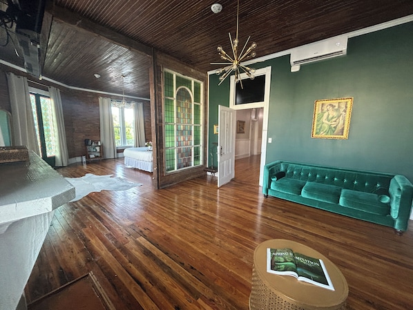 A spacious room with wooden flooring, a green sofa, a dining table, and a chandelier.