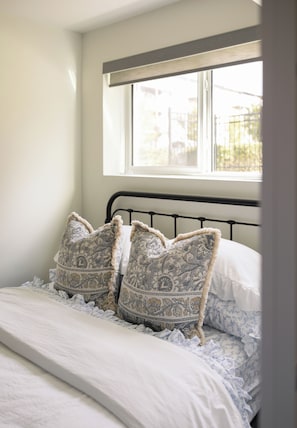 A bed with patterned pillows and a white comforter, a window above the bed, and a plain wall. - The Hugo Suite: Modern & Boutique (Winnipeg)