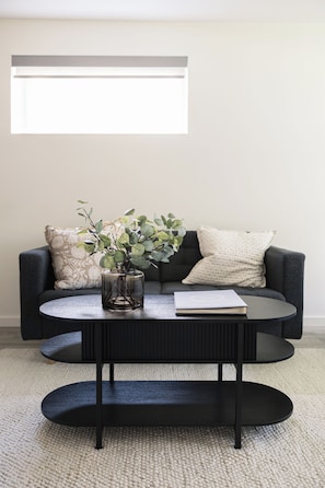 A modern living room with a round coffee table, a sofa with patterned cushions, and a window above. - The Hugo Suite: Modern & Boutique (Winnipeg)