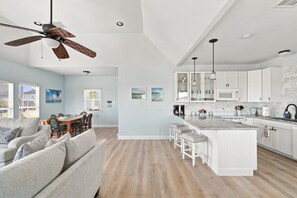 Un vaste espace de vie comprenant un coin repas avec une table et des chaises, une cuisine dotée de meubles blancs et d’un grand îlot central, ainsi qu’un ventilateur de plafond.