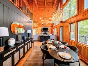 A wooden cabin interior with a dining area, kitchen, and a large window overlooking greenery. - Sunset Mtn Views + Sauna, Whirlpool Tub, Game Room (Bryson City)