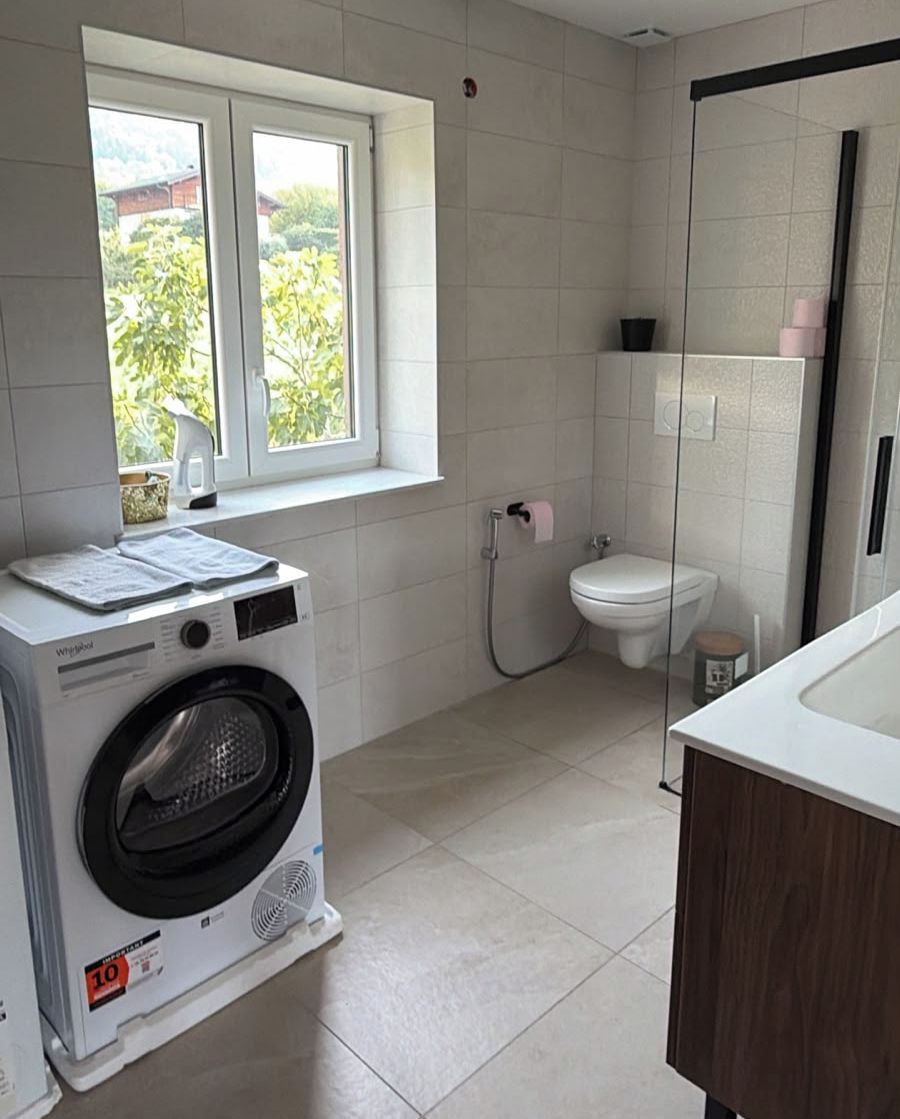 A compact bathroom with a washing machine, toilet, and a window providing natural light.