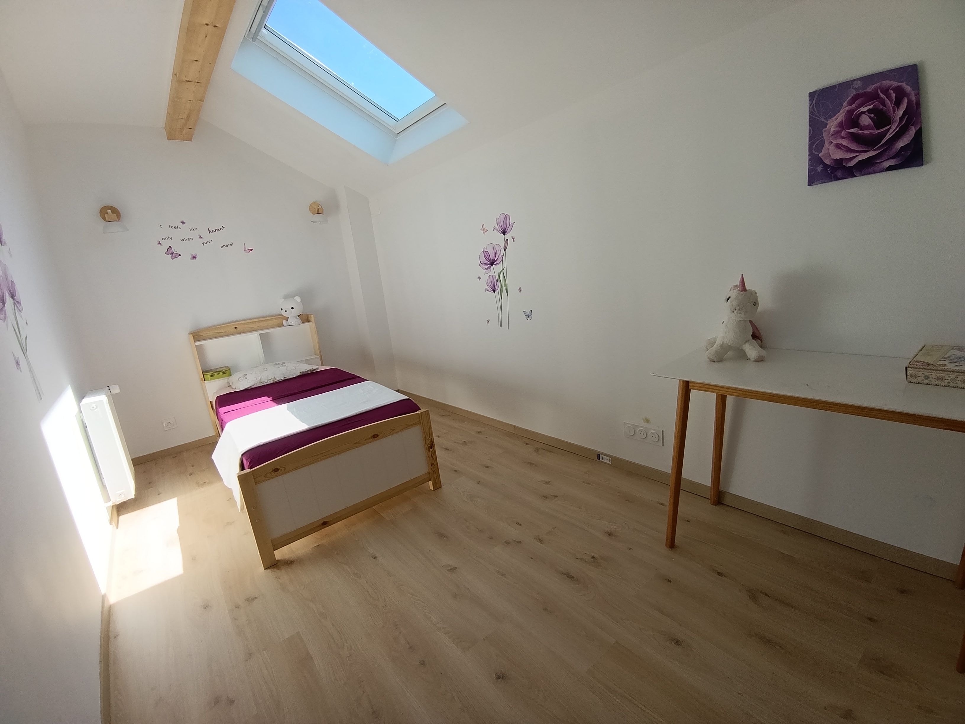 Une chambre avec un puits de lumière, un lit recouvert d’une couverture violette et blanche, une petite table sur laquelle est posé un jouet en peluche, et un mur décoré d’une rose violette.