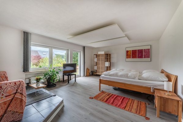 A modern bedroom with a bed, a TV on a stand, a bookshelf, and a window with a view of greenery.