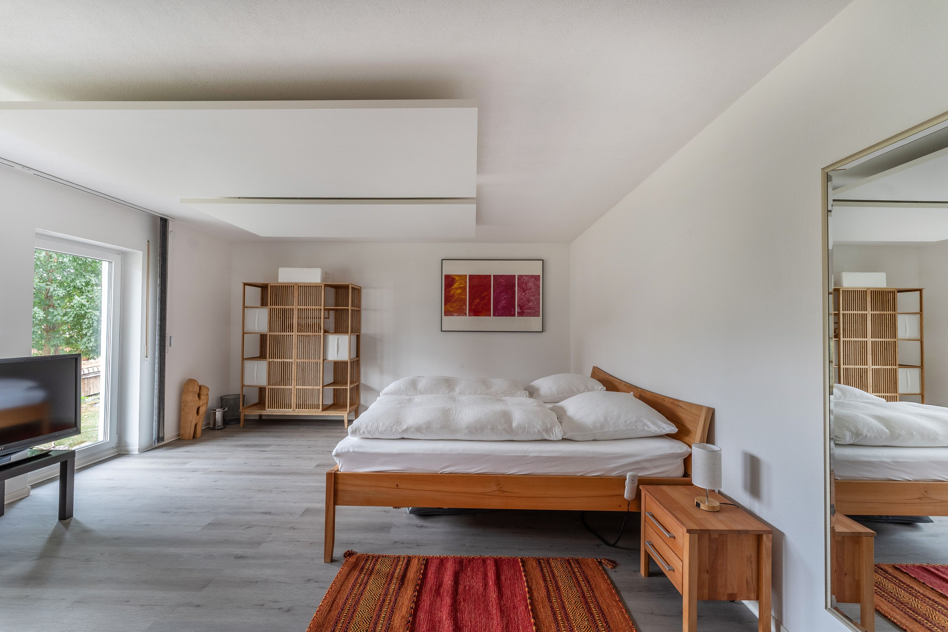 A modern bedroom with a wooden bed, a bedside table, a TV, and a large window.