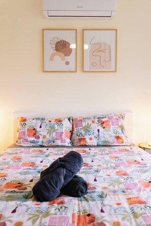 Ein Bett mit farbenfroher Blumendecke, zwei Kissen in ähnlichem Muster und zwei gerahmte Kunstwerke an der Wand darüber.