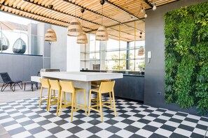 A modern interior with a checkered floor, a bar area with yellow chairs, hanging wicker lamps, and a green wall with plants. - Prime 1BR Flat in Las Condes Parking (Las Condes)
