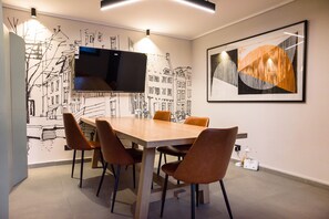 Ein moderner Essbereich mit einem langen Tisch, vier Stühlen, einem an der Wand montierten Flachbildfernseher und zwei Wandleuchten.