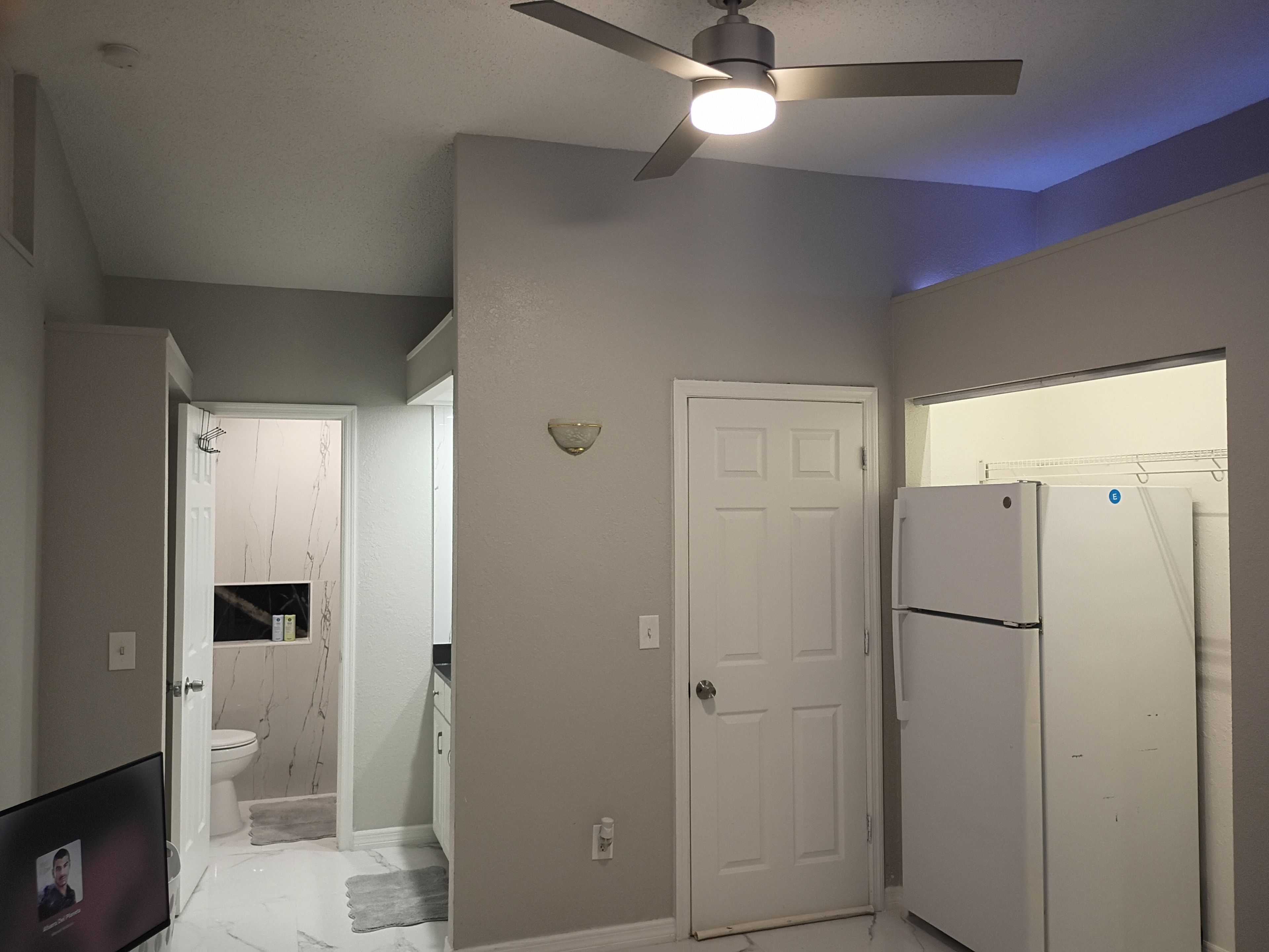 A compact kitchen with a refrigerator, a ceiling fan, and a door leading to another room.
