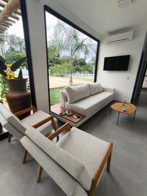 Een moderne woonkamer met een bank, twee fauteuils, een salontafel en een klein bijzettafeltje. Er is een groot raam met uitzicht op een tuin en palmbomen. Een flatscreen televisie is aan de muur bevestigd.