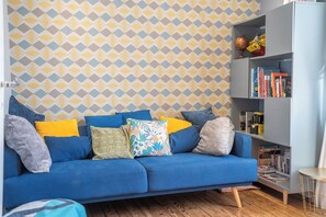 Ein Zimmer mit einem blauen Sofa, gemustertem Wandpapier, einem Bücherregal und einem kleinen runden Tisch.