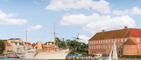 Un port où est amarré un grand voilier, ainsi qu’un plus petit bateau à voile, et un bâtiment historique surmonté d’un toit rouge.