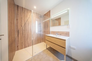Ein modernes Badezimmer mit einer Duschkabine aus Glas, Holzverkleidung, einem weißen Waschbecken und einem Spiegel mit integrierter Beleuchtung.