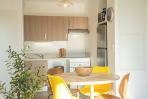 A compact kitchen with wooden cabinets, a stainless steel refrigerator, and a dining area with yellow chairs. - T2 neuf & lumineux Calme et plages à 5 min (Guidel)