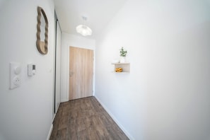 A hallway with a wooden door, a wall-mounted light fixture, and a small shelf with a plant. - T2 neuf & lumineux Calme et plages à 5 min (Guidel)