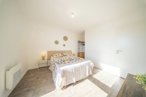 A bedroom with a bed, bedside tables, a radiator, and a door. - T2 neuf & lumineux Calme et plages à 5 min (Guidel)