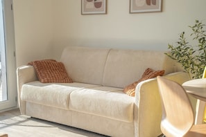 A beige corduroy sofa with two brown throw pillows, a wooden chair, and a potted plant. - T2 neuf & lumineux Calme et plages à 5 min (Guidel)