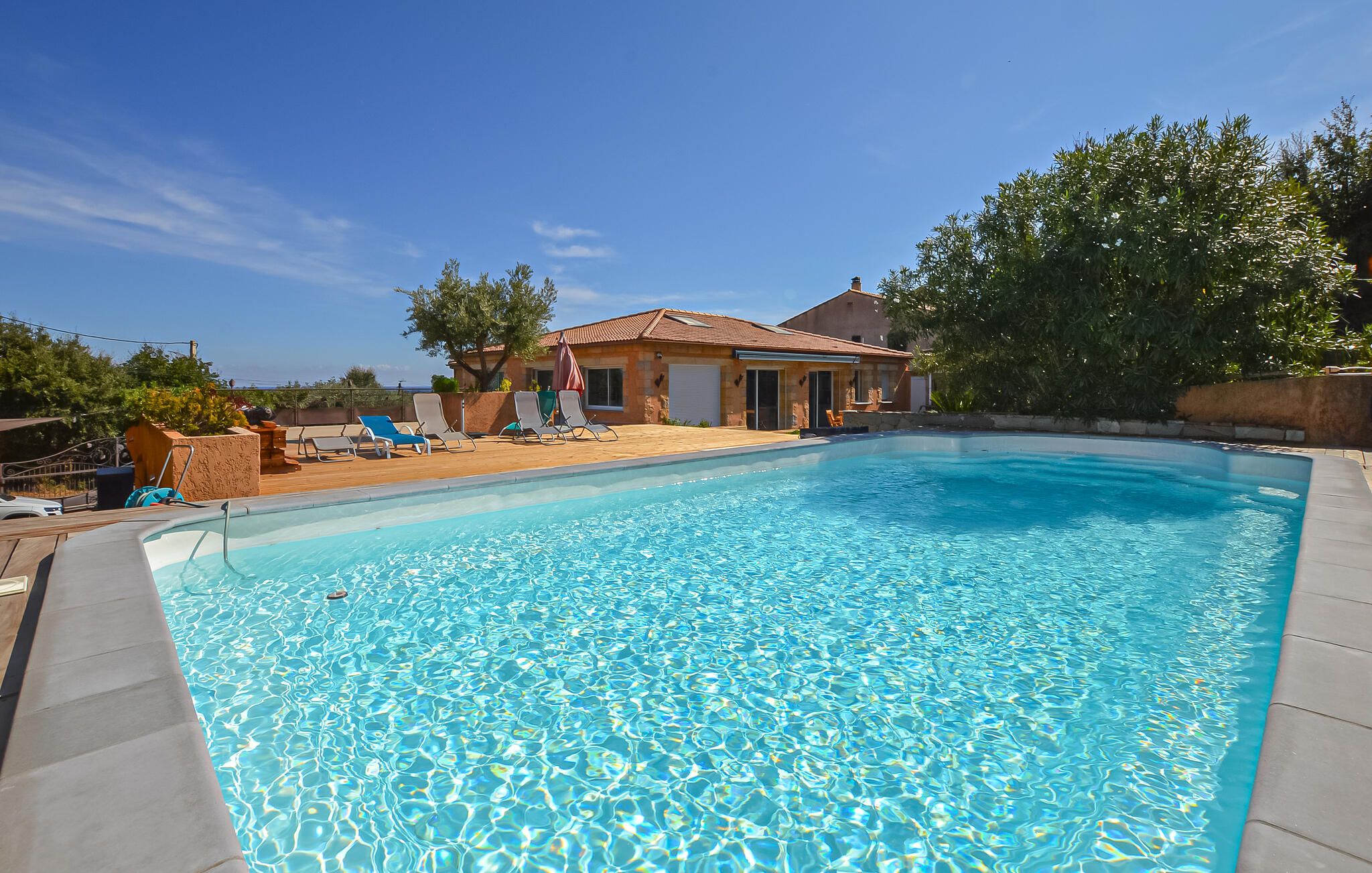 A swimming pool with clear blue water, surrounded by lounge chairs and a house with a tiled roof.