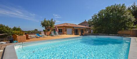 A swimming pool with clear blue water, surrounded by lounge chairs and a house with a tiled roof.
