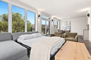 A modern hotel room with a large bed, a sofa, a wooden table, and a balcony with a view of trees. - Tunnel Vision Loft · 2BR by Trails & Bike Shop (Bella Vista)