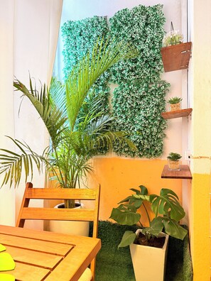 A room with a wooden table, chairs, a potted plant, a wall-mounted shelf with decorative items, and a green artificial wall. - Cozy & Secure 1BR Apartment City Center (Luanda)