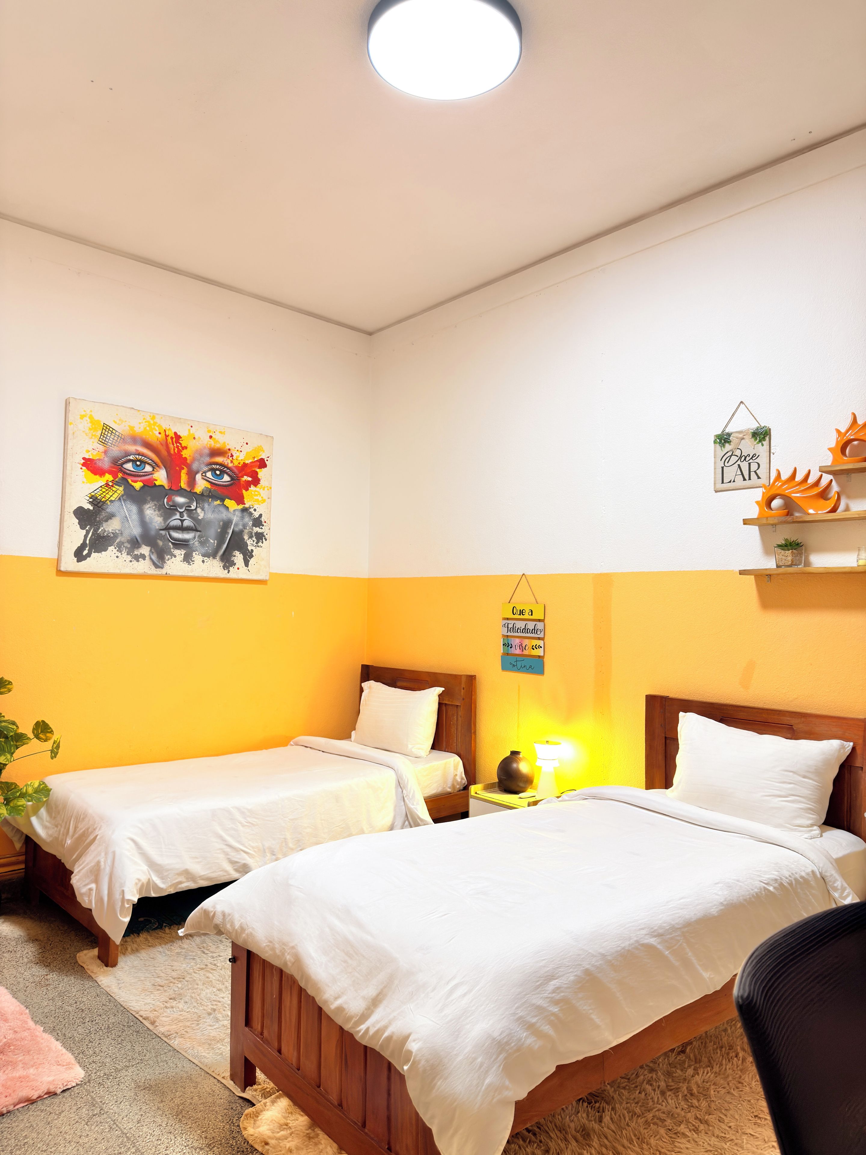 Habitación de hotel con dos camas, un cuadro en la pared, un pequeño estante con objetos decorativos y un cartel con texto.