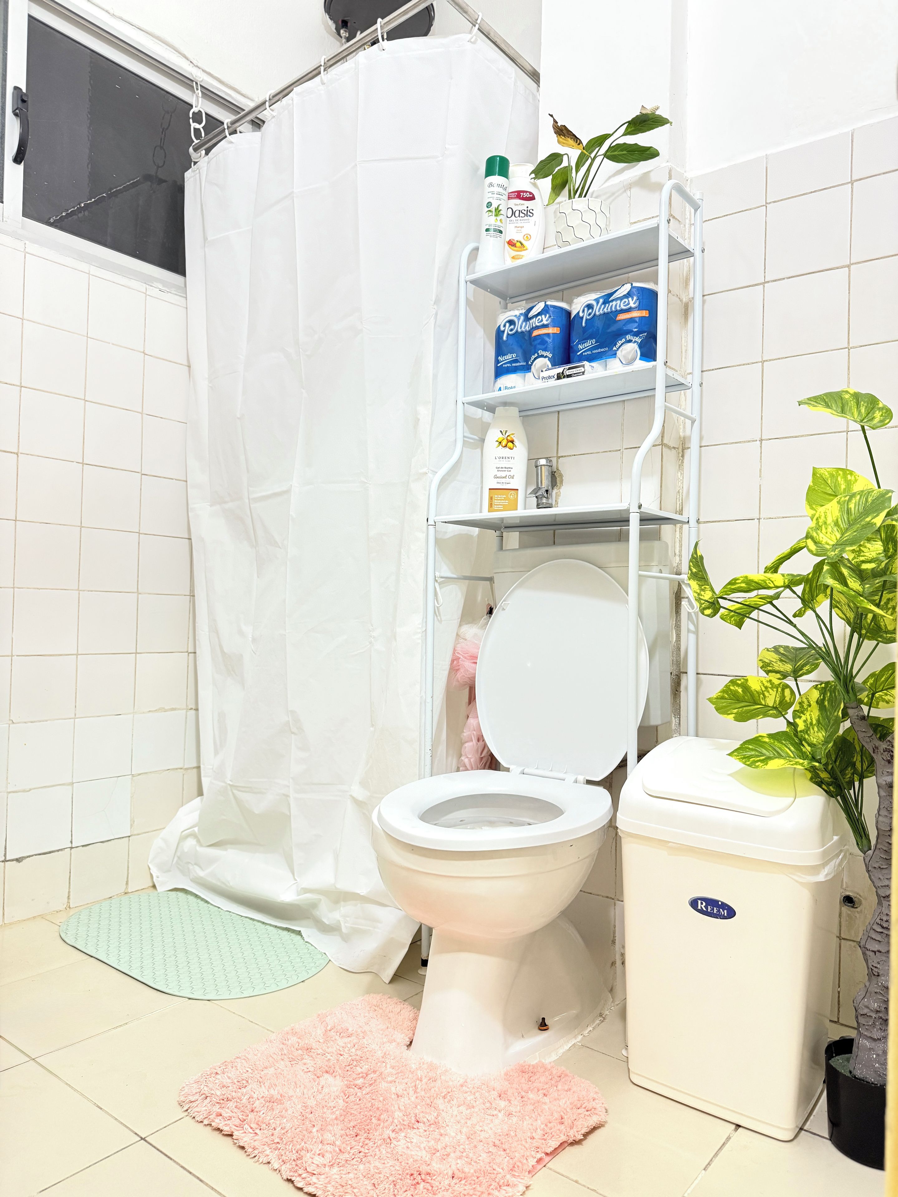 Un baño con inodoro, cortina de ducha, estante con artículos de aseo, alfombra rosa y una planta en maceta.