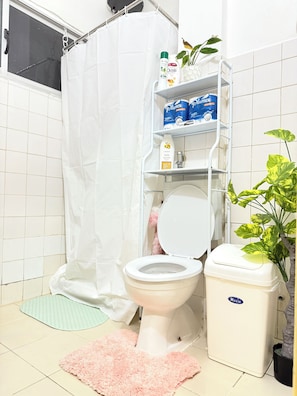 Un baño con inodoro, cortina de ducha, estante con artículos de aseo, alfombra rosa y una planta en maceta.