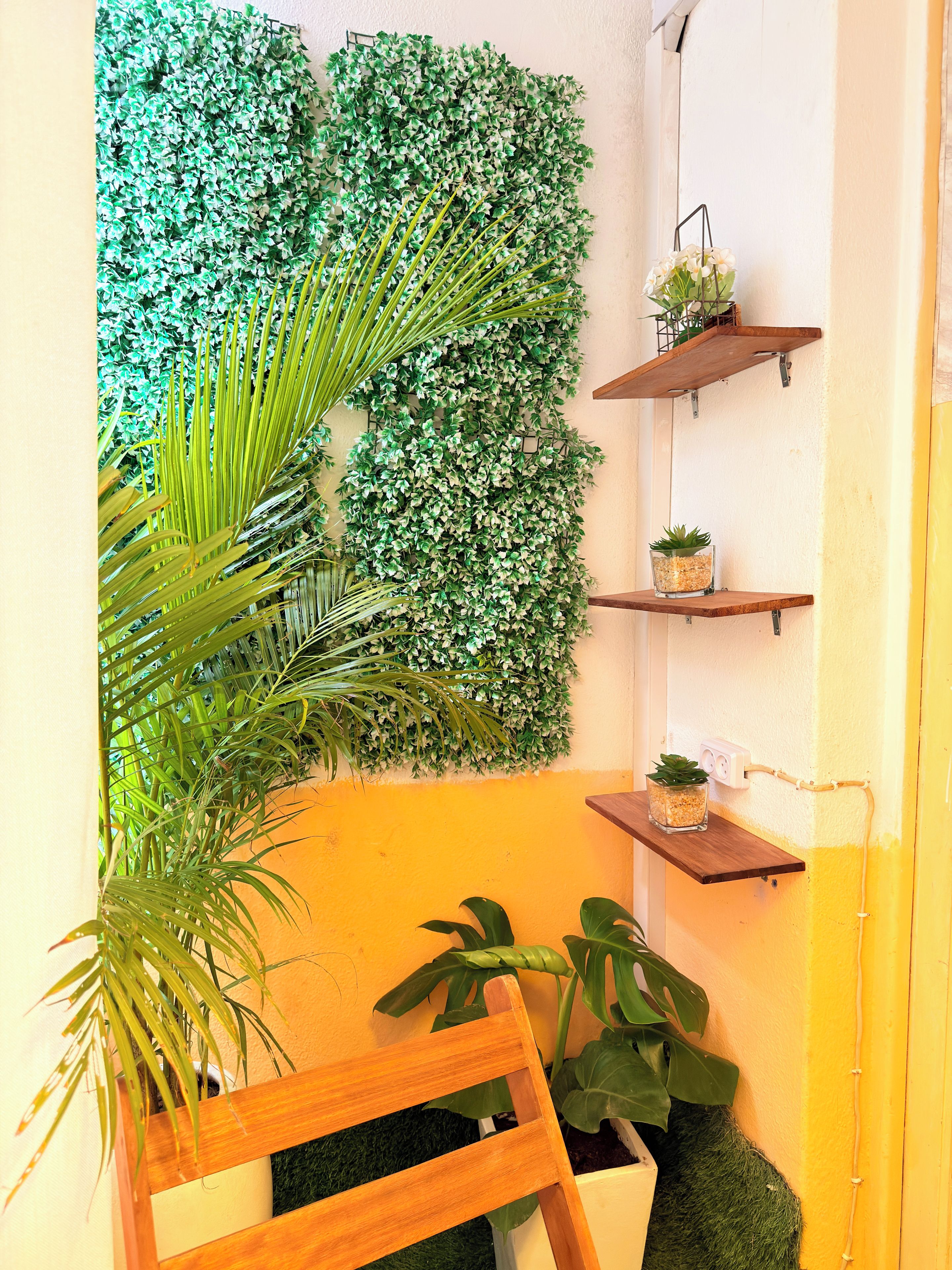 A room with a wall-mounted green foliage panel, a wooden chair, a small table, and two shelves with potted plants.