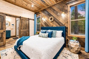 A bedroom with a large bed, wooden walls, a window with a view of trees, and a door leading to another room. - Games Inside & Out | Hot Tub | Sleeps 11 (Broken Bow)
