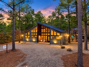 A modern cabin with large windows, surrounded by trees, and a gravel driveway. - Games Inside & Out | Hot Tub | Sleeps 11 (Broken Bow)