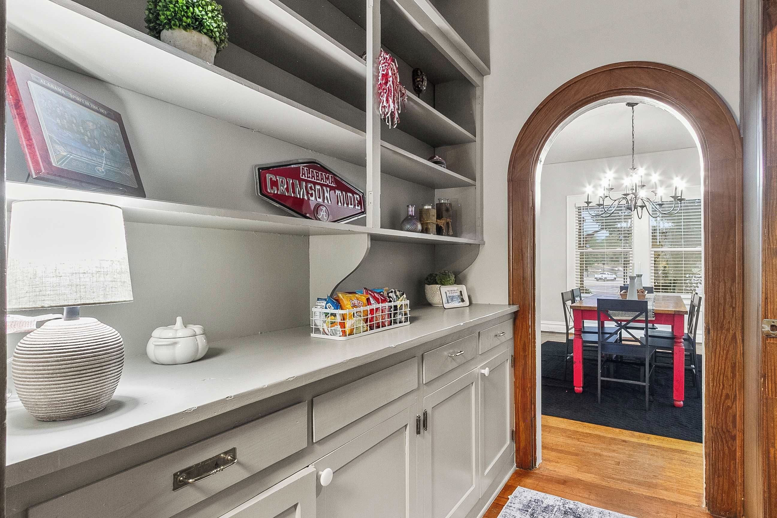 A kitchen with a countertop, cabinets, a potted plant, a sign, and a dining area with a table and chairs.
