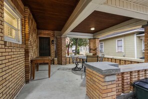 A covered patio area with brick walls, a wooden table, and a seating arrangement. - Tide & True | 5BR Walk to UA | Prime Location (Tuscaloosa)