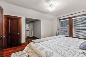 A bedroom with a wooden door, a bed with a patterned bedspread, a ceiling fan, and a window with blinds. - Tide & True | 5BR Walk to UA | Prime Location (Tuscaloosa)