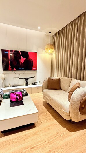 Ein modernes Wohnzimmer mit einem Flachbildfernseher, einem weißen Couchtisch, einem beigen Sofa und einer dekorativen Lampe.