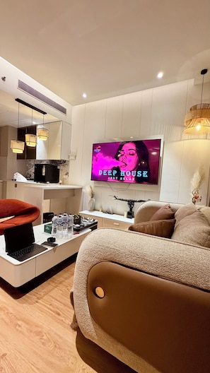 A modern living room with a flat-screen TV, a sofa, a coffee table, and a dining area with a refrigerator. - Luxury Paradise Studio Apartment | Elegant & Peaceful Stay (Gurugram)