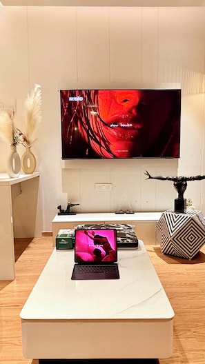 A modern living room with a flat-screen TV mounted on the wall, a laptop on a white coffee table, and decorative items including a vase and a geometric sculpture. - Luxury Paradise Studio Apartment | Elegant & Peaceful Stay (Gurugram)
