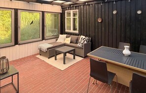 Eine überdachte Terrasse mit einer Couch, einem Tisch und Stühlen, großen Fenstern und einem Ziegelboden.