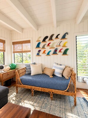 A room with a rattan sofa, a wooden desk, and a wall-mounted surfboard display. - Poipu Ocean View Beach House  - Sleeps 6 (Koloa)