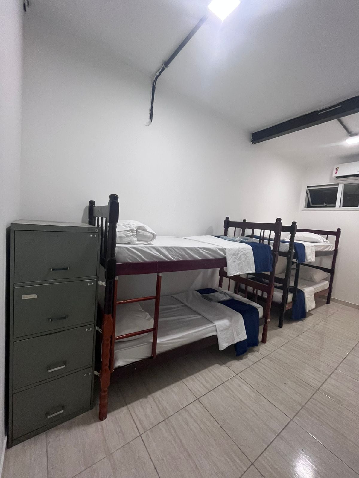 A dormitory room with bunk beds, a filing cabinet, and a window.