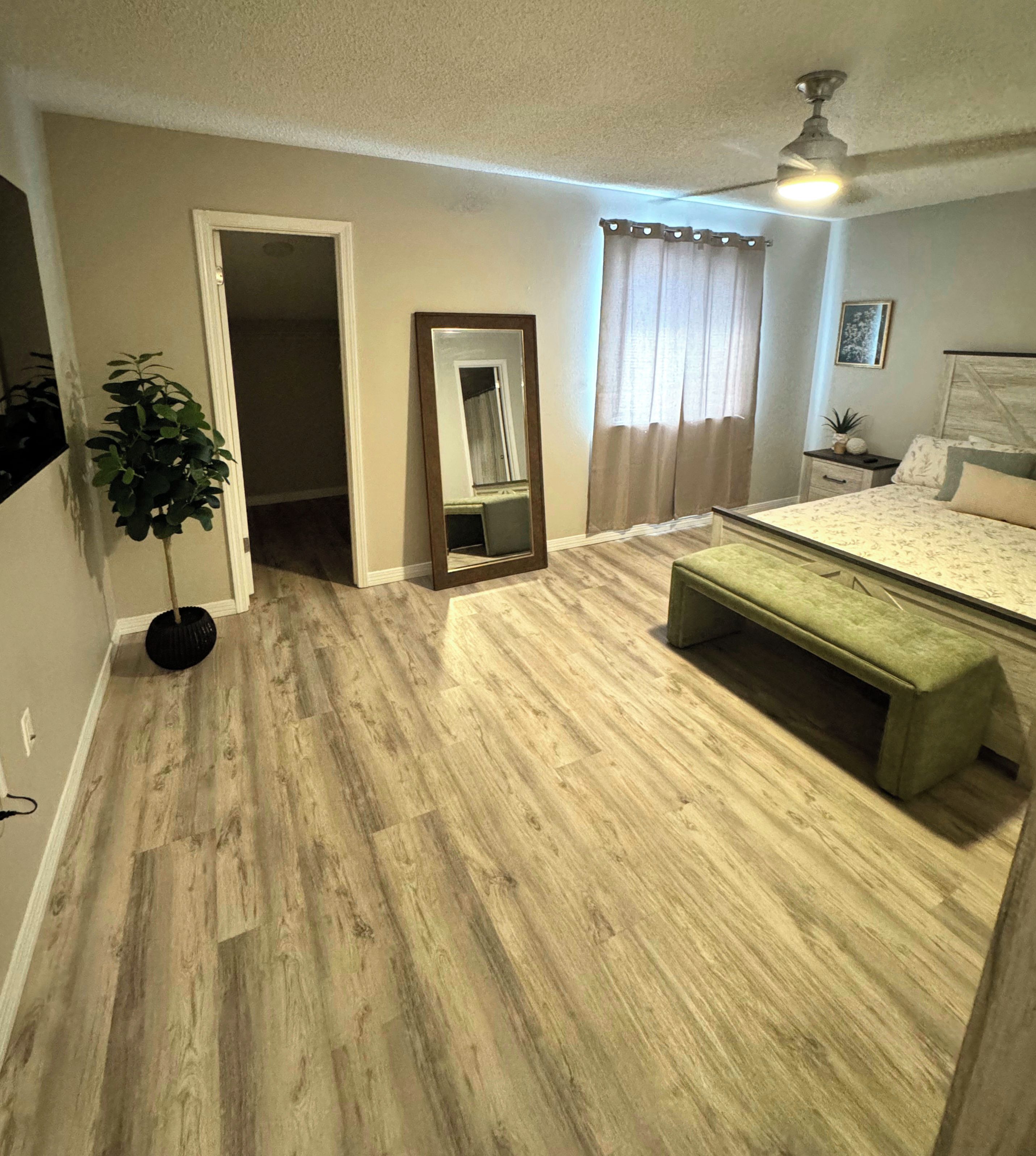 Un dormitorio con piso de madera, una cama con un banco tapizado en verde, una mesita de noche con una planta y un ventilador de techo.