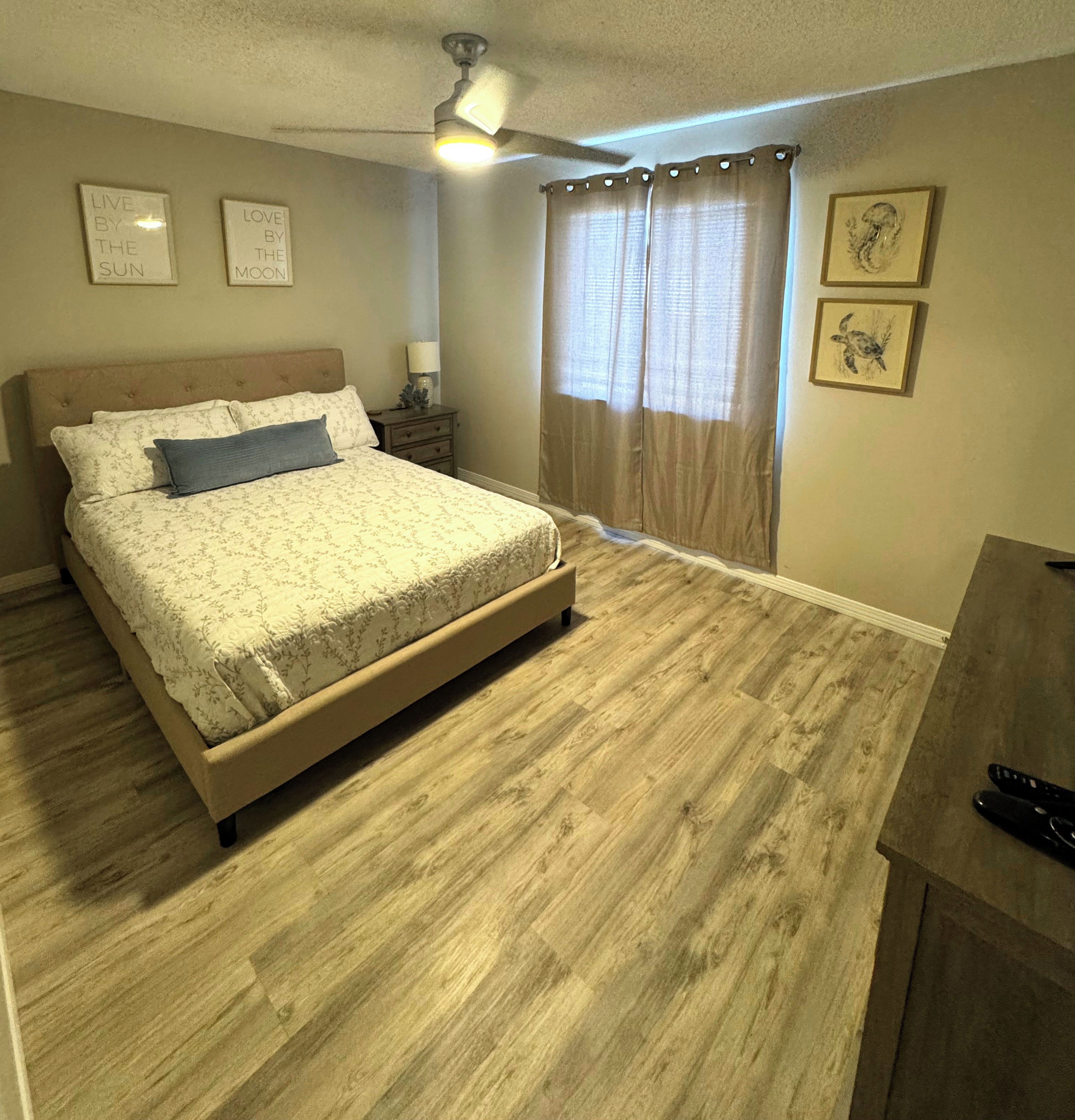 Un dormitorio con cama, mesita de noche, lámpara, ventilador de techo y ventana con cortinas.