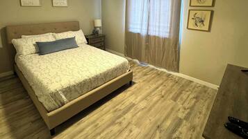 Un dormitorio con cama, mesita de noche, lámpara, ventilador de techo y ventana con cortinas.