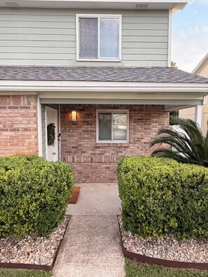 A residential house with a brick facade, a front door, a small window, and two well-maintained bushes.
