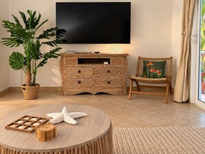 Ein Wohnzimmer mit Flachbildfernseher, einem Korbstuhl mit Blumenkissen, einem Holztisch mit Seestern und Holzbausteinen und einer großen Pflanze in einem gewebten Korb.