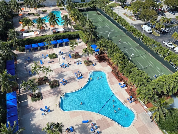 Una piscina con sillones de descanso y sombrillas, rodeada de palmeras y una cancha de tenis.